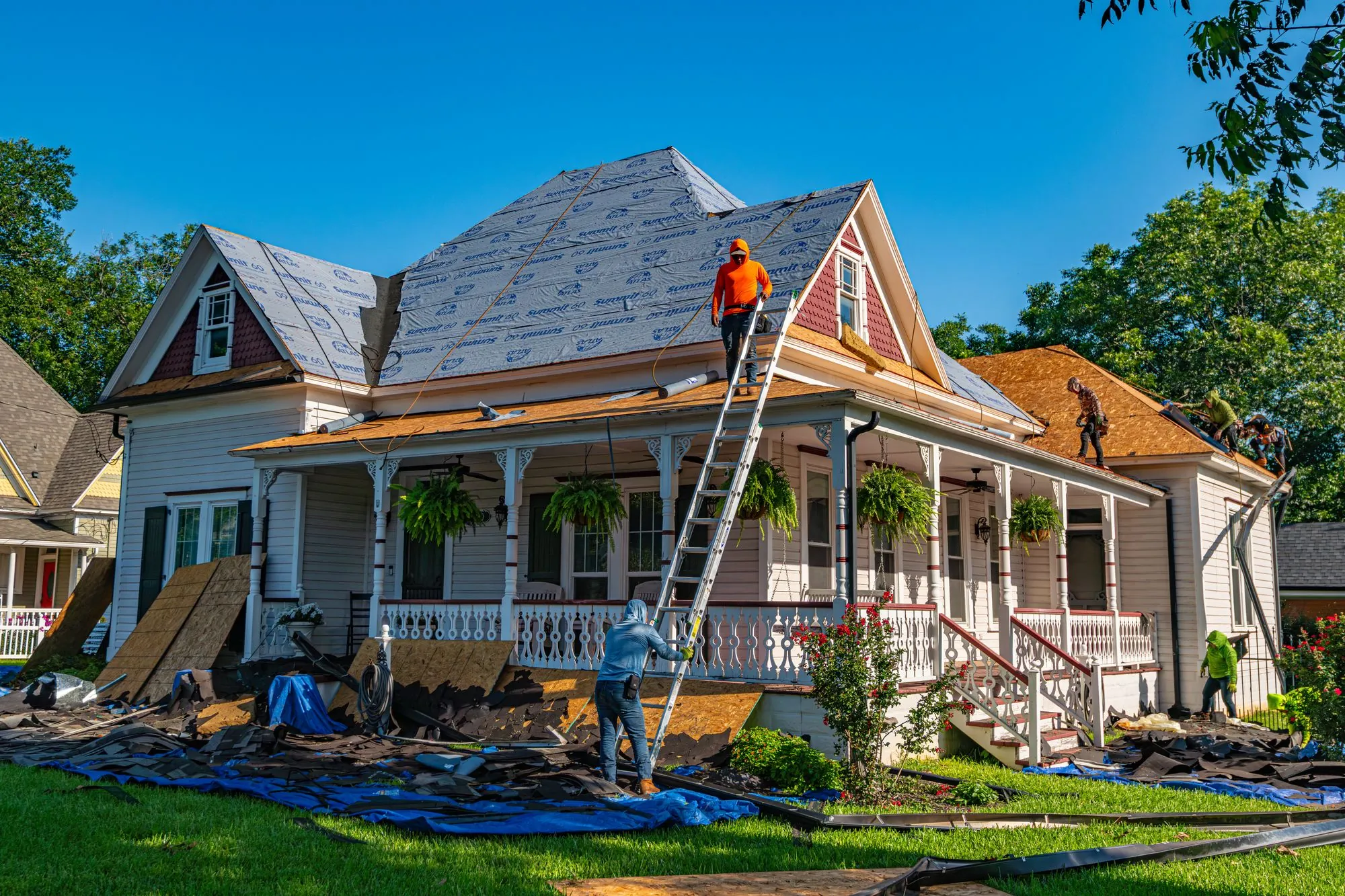 A roofing crew re-covering a house, ladders up and the beds sheeted over