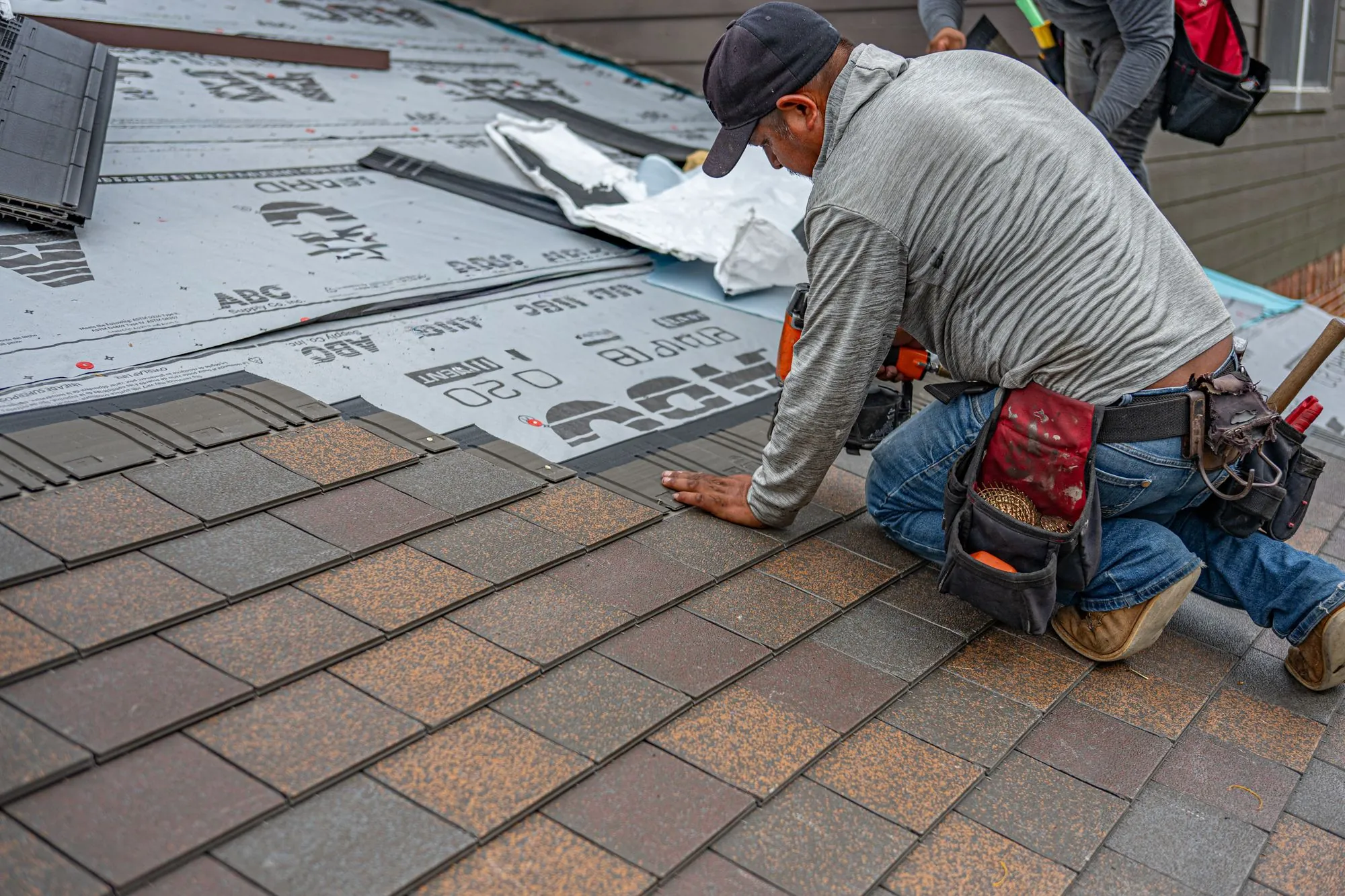 A roofer kneeling on a residential roof, setting a course of asphalt shingles